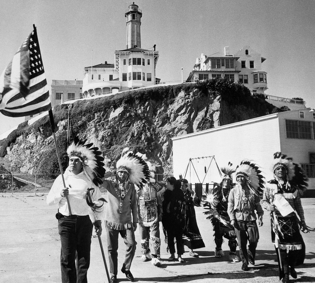 a group of Native Americans holding an American flag on a beach with buildings in the background (alcatraz)