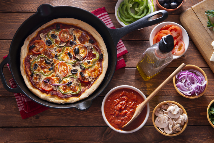 pizza in a skillet alongside bowls of ingredients like sauce and veggies