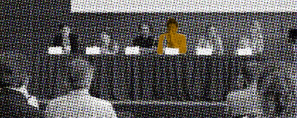 several people sitting on a stage facing an audience, with one person in particular highlighted to stand out