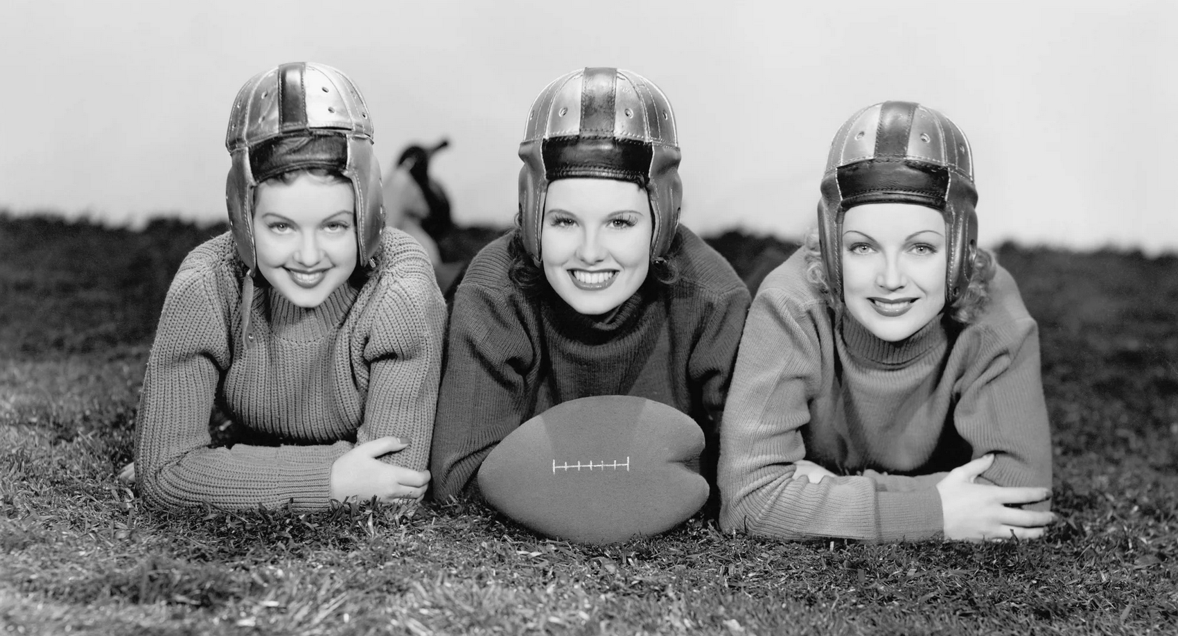 three smiling women posing on the grass with a football